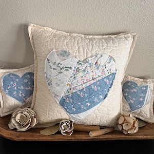 Puede incluir: Una almohada acolchada de color crema con un diseño de corazón de retazos, flanqueada por dos almohadas más pequeñas en forma de corazón. Los diseños de corazón presentan patrones florales y azules. Flores decorativas de madera y un cuenco de madera completan la exhibición.