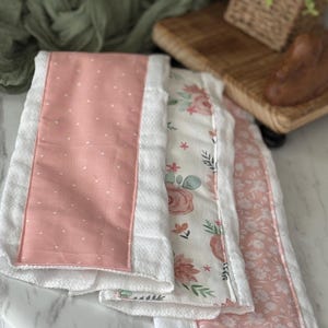Puede incluir: Tres toallas de mano decorativas blancas y rosas. Una tiene un diseño de lunares rosas, otra un estampado floral y la tercera un pequeño estampado floral. Las toallas están dobladas y apiladas sobre una superficie blanca.
