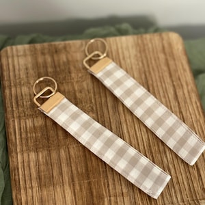 Puede incluir: Dos llaveros con un estampado de cuadros vichy beige y blanco. Cada llavero tiene un anillo dorado y un detalle de cuero marrón claro. Los llaveros se muestran sobre una tabla de cortar de madera.