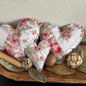 Puede incluir: Tres decoraciones de tela en forma de corazón con estampados florales en rosa, rojo y blanco, dispuestas en un cuenco de madera con elementos decorativos. Los corazones tienen un diseño de patchwork y están rellenos de relleno.