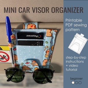Op de afbeelding: Een mini-auto-zonneklep-organizer gemaakt van lichtblauwe stof met een patroon van cartoonberen en kersen. De organizer heeft vakken voor kaarten, een penhouder en een zonnebril. De tekst "MINI CAR VISOR ORGANIZER" staat bovenaan.