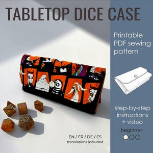 Puede incluir: Un estuche rectangular de tela para dados con un estampado negro y naranja de personajes de Pesadilla antes de Navidad, cerrado con un broche plateado. Cinco dados multifacéticos están dispuestos delante. Texto: TABLETOP DICE CASE.