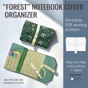 May include: A Forest notebook cover organizer featuring a green fabric with an acorn and leaf print, a cream strap with metal snaps, and an interior with a yellow floral lining, pen slots, and a pocket. Includes text: FOREST NOTEBOOK COVER ORGANIZER.