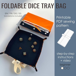 May include: A foldable dice tray bag featuring a black interior and a bright orange exterior. The tray is shown in an open, square configuration with seven polyhedral dice inside. A separate closed pouch section is visible with a zipper and a pencil.