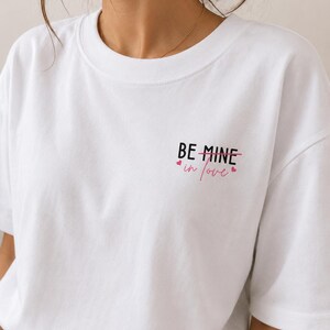 May include: White t-shirt featuring the text "BE MINE" crossed out and replaced with "in love" in pink, accompanied by small pink heart graphics. The top has a crew neck and short sleeves.