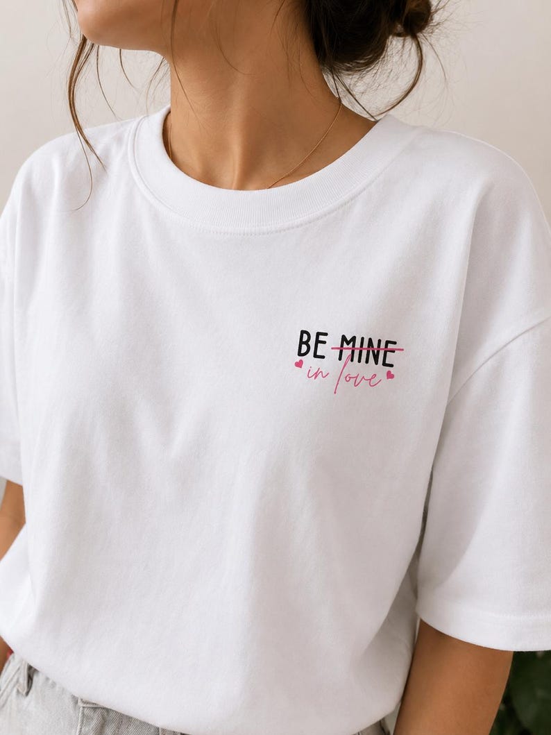 May include: White t-shirt featuring the text "BE MINE" crossed out and replaced with "in love" in pink, accompanied by small pink heart graphics. The top has a crew neck and short sleeves.