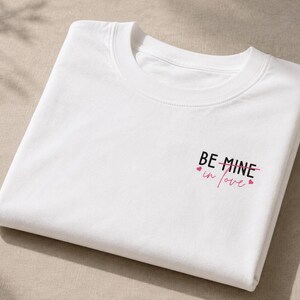 May include: A folded white crew neck t-shirt. The text "BE MINE in love" is printed in black and pink, with small pink hearts. The shirt is made of a soft material. The design is on the front.