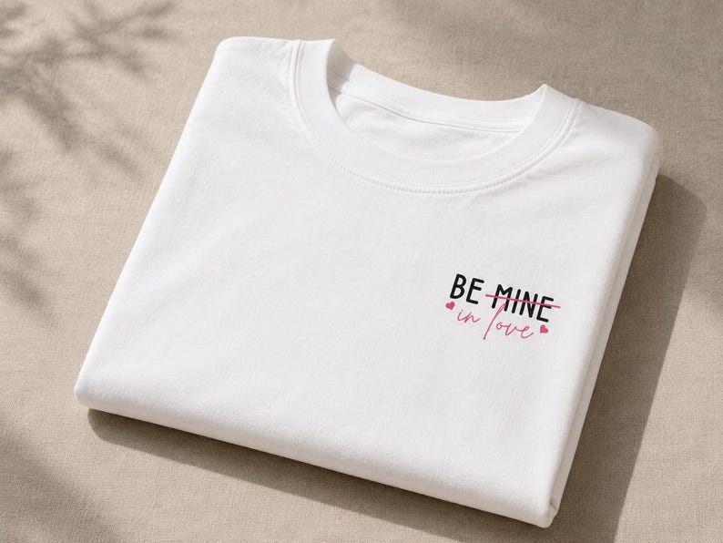 May include: A folded white crew neck t-shirt. The text "BE MINE in love" is printed in black and pink, with small pink hearts. The shirt is made of a soft material. The design is on the front.