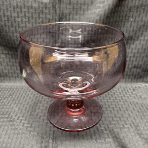 May include: A vintage, stemmed glass in a soft pink hue. The bowl-shaped top sits atop a short, rounded stem and a circular base. The glass has a smooth, reflective surface, and the overall design is simple and elegant.