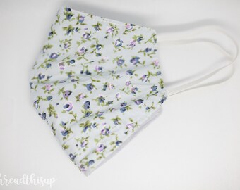 May include: A white face mask with a floral pattern featuring small blue and pink flowers. The mask has elastic ear loops.
