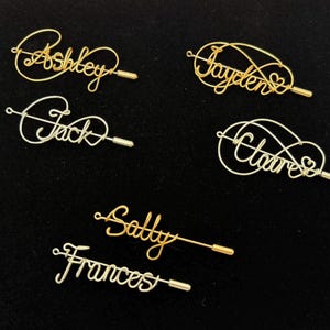 May include: A selection of personalised wire name pins in gold and silver. Each pin features a different name in a cursive script, including "Ashley", "Jayden", "Jack", "Claire", "Sally", and "Frances". The pins are displayed on a black surface.