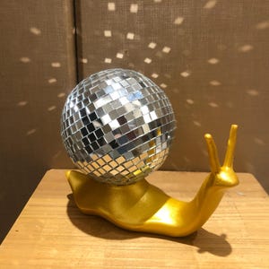 May include: A decorative ornament of a golden snail with a mirrored disco ball shell. The snail has two upright antennae. The disco ball is made of small, reflective squares. The piece is designed to catch and reflect light.