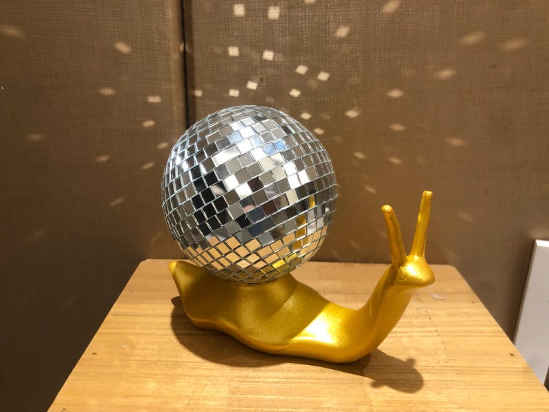 Puede incluir: Una pieza decorativa caprichosa que presenta una bola de discoteca plateada con espejos sobre una figura de caracol dorada. El caracol tiene dos antenas verticales. La bola de discoteca refleja la luz.