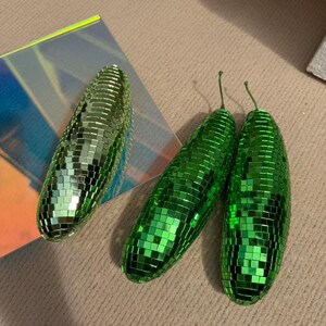 May include: Three green disco ball-style ornaments. Two are elongated, with small stems, and one is oval-shaped. The ornaments are covered in small, reflective squares, creating a shimmering effect. They are placed on a neutral-toned surface.