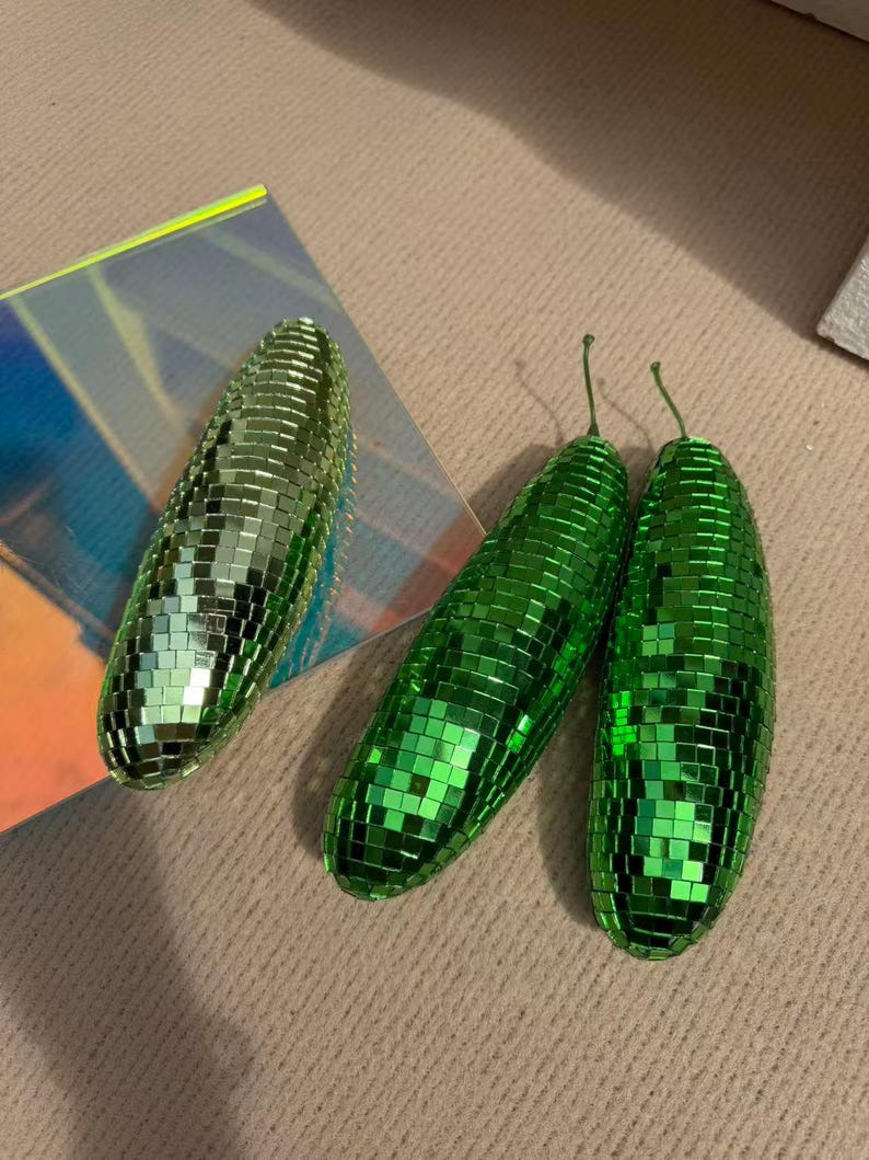May include: Three green disco ball-style ornaments. Two are elongated, with small stems, and one is oval-shaped. The ornaments are covered in small, reflective squares, creating a shimmering effect. They are placed on a neutral-toned surface.
