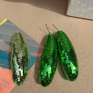 May include: Three green disco ball-style ornaments. Two are upright, and one is lying on its side. The ornaments are covered in small, reflective squares. The ornaments have a stem at the top.