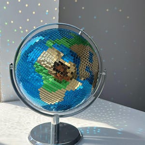 Könnte beinhalten: Ein dekorativer Globus mit verspiegelter Oberfläche, der Licht reflektiert. Der Globus hat blaue, grüne und goldfarbene Spiegelfliesen, die auf einem silberfarbenen Metallständer montiert sind. Die Kontinente sind goldfarben umrandet. Ein einzigartiger Einrichtungsgegenstand.