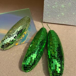 May include: Three green disco ball-style decorative objects. Two are side-by-side, and one is positioned separately. The objects have a reflective, mosaic-like surface, and a small green stem. The objects are on a neutral-coloured surface.