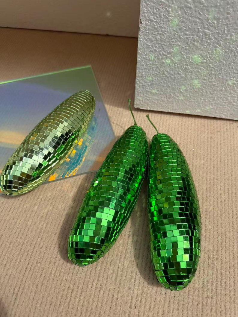 May include: Three green disco ball-style decorative objects. Two are side-by-side, and one is positioned separately. The objects have a reflective, mosaic-like surface, and a small green stem. The objects are on a neutral-coloured surface.