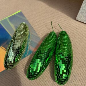 May include: Three green disco ball-style ornaments. Two are elongated, with small green mirrored tiles. The third is a similar shape, but slightly smaller. The ornaments have a stem-like detail at the top. They are on a neutral surface.