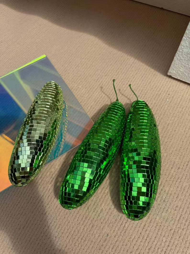 May include: Three green disco ball-style ornaments. Two are elongated, with small green mirrored tiles. The third is a similar shape, but slightly smaller. The ornaments have a stem-like detail at the top. They are on a neutral surface.