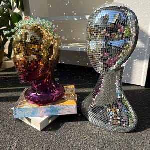 Puede incluir: Dos cabezas de maniquí decorativas cubiertas de azulejos espejados. Una cabeza es dorada y morada, adornada con una corona de cuentas. La otra es plateada. Ambas se exhiben sobre una superficie oscura, con libros debajo de la cabeza dorada.