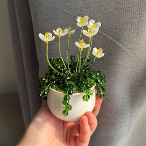 May include: A miniature white ceramic pot containing beaded flowers. The flowers feature white petals with yellow centres and green stems. The pot is held in a hand against a grey backdrop. The beaded foliage is a deep green.