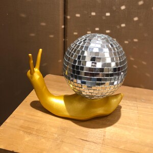 Puede incluir: Una escultura caprichosa de un caracol dorado con una concha de bola de discoteca plateada. El caracol est&aacute; colocado sobre una superficie de madera, con la luz reflejada en la bola de discoteca. Un art&iacute;culo decorativo &uacute;nico.