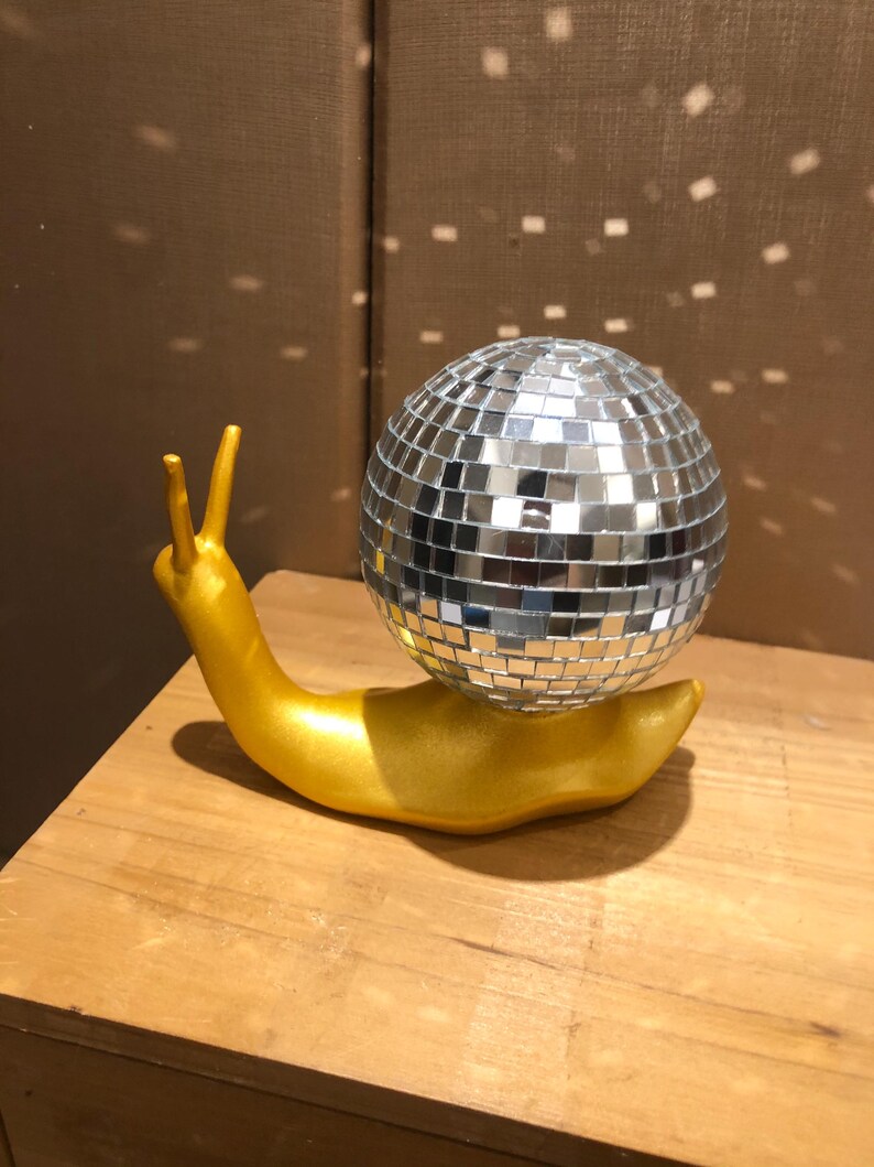 Puede incluir: Una escultura caprichosa de un caracol dorado con una concha de bola de discoteca plateada. El caracol est&aacute; colocado sobre una superficie de madera, con la luz reflejada en la bola de discoteca. Un art&iacute;culo decorativo &uacute;nico.