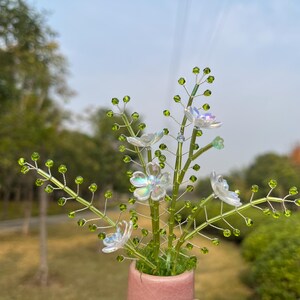 Könnte beinhalten: Ein handgefertigtes Blumenarrangement mit zarten, irisierenden Blüten und grünen Perlenstielen. Die Stiele sind in einem kleinen, rosa Keramiktopf vor einem verschwommenen Hintergrund im Freien angeordnet.