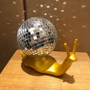 Puede incluir: Una escultura caprichosa de un caracol dorado con una concha de bola de discoteca espejada. El caracol est&aacute; sobre una superficie de madera, y la bola de discoteca refleja la luz, creando un efecto brillante. Un art&iacute;culo decorativo &uacute;nico.