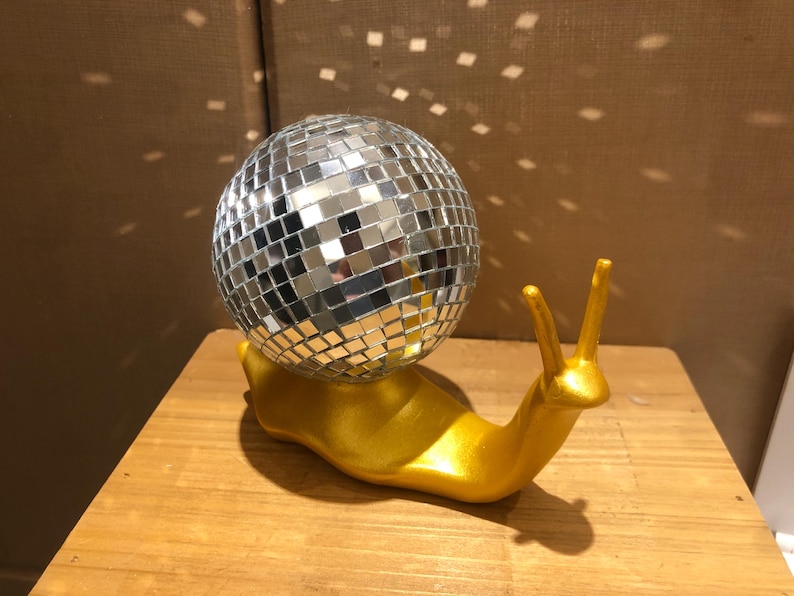 Puede incluir: Una escultura caprichosa de un caracol dorado con una concha de bola de discoteca espejada. El caracol est&aacute; sobre una superficie de madera, y la bola de discoteca refleja la luz, creando un efecto brillante. Un art&iacute;culo decorativo &uacute;nico.