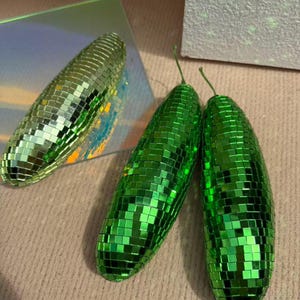 May include: Three decorative, oblong objects covered in small, reflective green squares, resembling disco balls. One is a lighter green, while the other two are a vibrant, darker green. The objects are arranged on a neutral, textured surface.