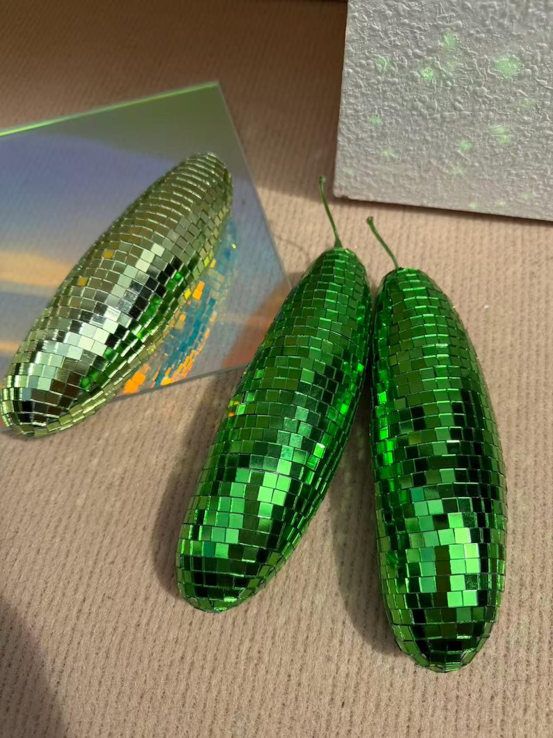 May include: Three decorative, oblong objects covered in small, reflective green squares, resembling disco balls. One is a lighter green, while the other two are a vibrant, darker green. The objects are arranged on a neutral, textured surface.