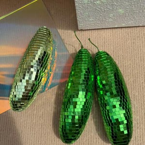 May include: Three disco ball-style ornaments shaped like elongated ovals. Two are bright green, and one is a lighter, more iridescent green. Each is covered in small, reflective squares, and the green ones have a small green stem.