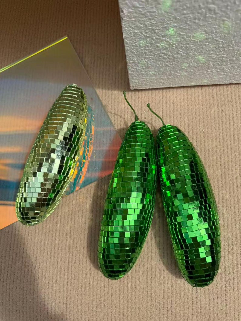 May include: Three disco ball-style ornaments shaped like elongated ovals. Two are bright green, and one is a lighter, more iridescent green. Each is covered in small, reflective squares, and the green ones have a small green stem.