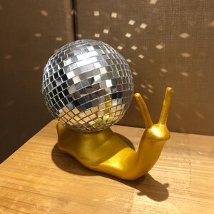 Puede incluir: Una original figura decorativa de un caracol dorado con una bola de discoteca espejada. El cuerpo del caracol es dorado y la bola de discoteca refleja la luz. Este objeto decorativo a&ntilde;ade un toque divertido y brillante a cualquier espacio.