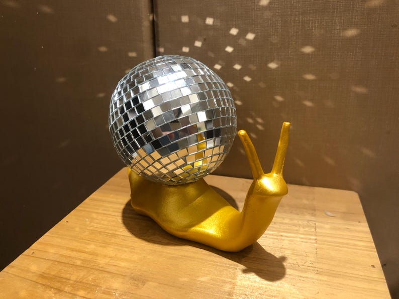 Puede incluir: Una original figura decorativa de un caracol dorado con una bola de discoteca espejada. El cuerpo del caracol es dorado y la bola de discoteca refleja la luz. Este objeto decorativo a&ntilde;ade un toque divertido y brillante a cualquier espacio.