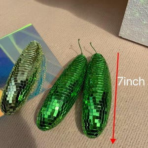 May include: Three green, disco ball-style decorative objects. Two are elongated, measuring 18 cm in length, and one is oval-shaped. The objects are covered in small, reflective squares, creating a shimmering effect. The objects have a stem.
