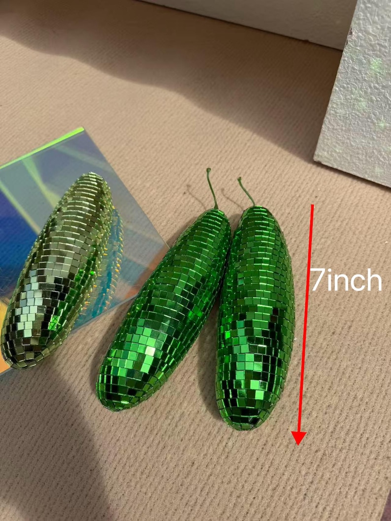 May include: Three green, disco ball-style decorative objects. Two are elongated, measuring 18 cm in length, and one is oval-shaped. The objects are covered in small, reflective squares, creating a shimmering effect. The objects have a stem.