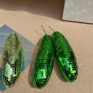 May include: Three green, disco ball-style ornaments. Two are upright, and one is on its side. Each has a small green stem. The ornaments are covered in small, reflective squares, creating a shimmering effect. The background is a light beige colour.