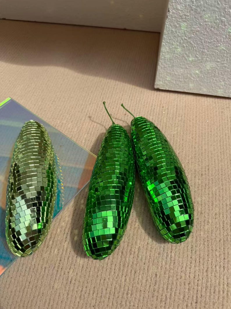 May include: Three green, disco ball-style ornaments. Two are upright, and one is on its side. Each has a small green stem. The ornaments are covered in small, reflective squares, creating a shimmering effect. The background is a light beige colour.