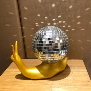 Puede incluir: Una escultura caprichosa de un caracol dorado que sostiene una bola de discoteca. El caracol es dorado y la bola est&aacute; cubierta de peque&ntilde;os cuadrados espejados. La escultura est&aacute; sobre una superficie de madera.