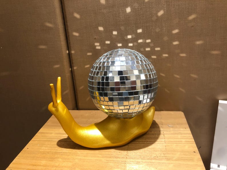 Puede incluir: Una escultura caprichosa de un caracol dorado que sostiene una bola de discoteca. El caracol es dorado y la bola est&aacute; cubierta de peque&ntilde;os cuadrados espejados. La escultura est&aacute; sobre una superficie de madera.