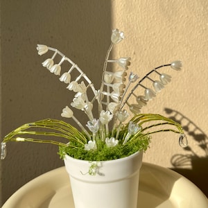 Pode incluir: Um pequeno vaso de cerâmica branco cheio de flores artificiais de lírio-do-vale. As flores são brancas com detalhes transparentes, e os caules são prateados e verdes. O vaso está numa bandeja amarelo claro.