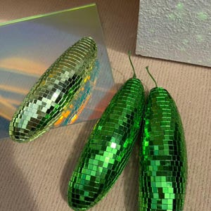 May include: Three green disco ball ornaments with a mirrored surface. The ornaments are oblong, with a small green stem. One is on a reflective surface, the others on a neutral background. Each is covered in small, reflective squares.