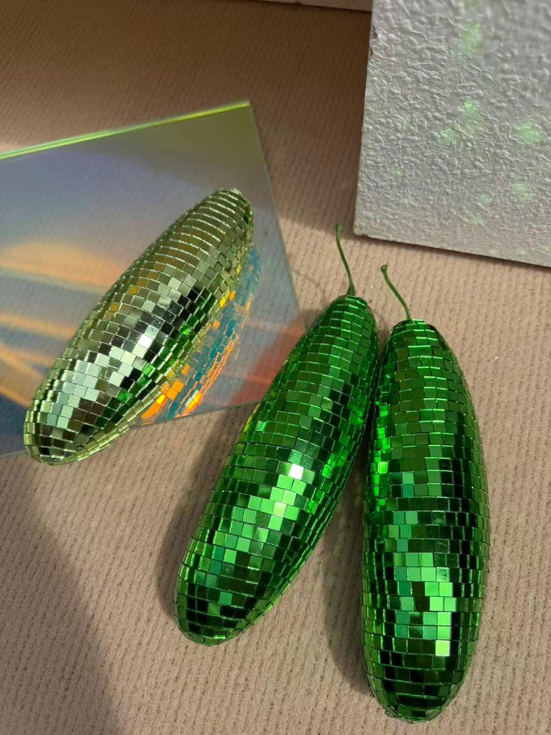 May include: Three green disco ball ornaments with a mirrored surface. The ornaments are oblong, with a small green stem. One is on a reflective surface, the others on a neutral background. Each is covered in small, reflective squares.