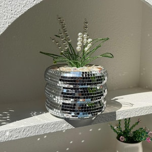 May include: A silver mosaic planter with a layered design, containing faux greenery and pearl-like accents. The planter is on a white shelf, creating a modern display. The planter is approximately 15 cm in diameter.