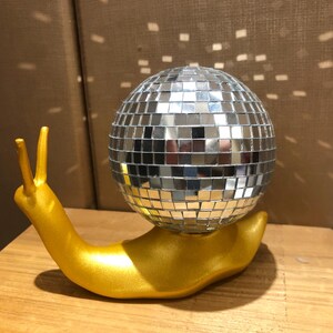 Puede incluir: Una escultura caprichosa de un caracol dorado con una concha de bola de discoteca espejada. El caracol est&aacute; posado sobre una superficie de madera, reflejando la luz. Un art&iacute;culo decorativo &uacute;nico.
