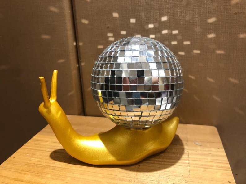 Puede incluir: Una escultura caprichosa de un caracol dorado con una concha de bola de discoteca espejada. El caracol est&aacute; posado sobre una superficie de madera, reflejando la luz. Un art&iacute;culo decorativo &uacute;nico.